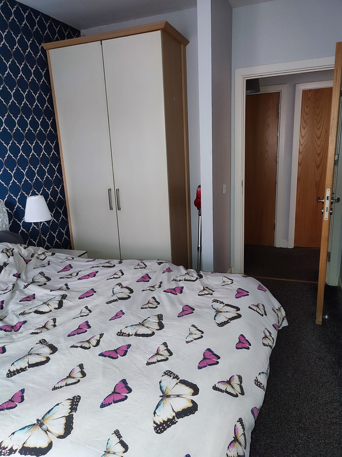A spacious bedroom features a bed adorned with a butterfly-patterned duvet. A large wardrobe is positioned against a patterned blue wall, and an open door leads to a hallway with additional doors visible.