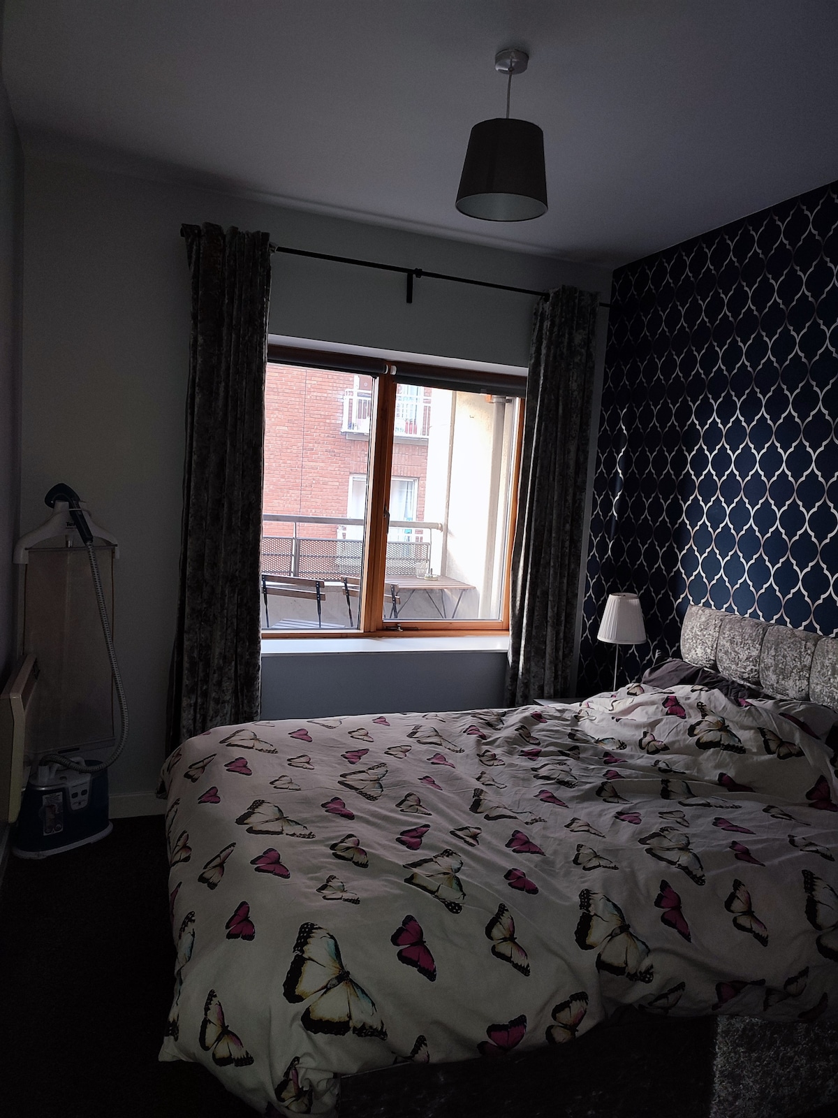 A cozy bedroom is furnished with a spacious bed covered in a butterfly-patterned duvet. A large window, dressed with patterned curtains, fills the room with natural light. A bedside lamp and clothing steamer are also visible, contributing to the functional space.