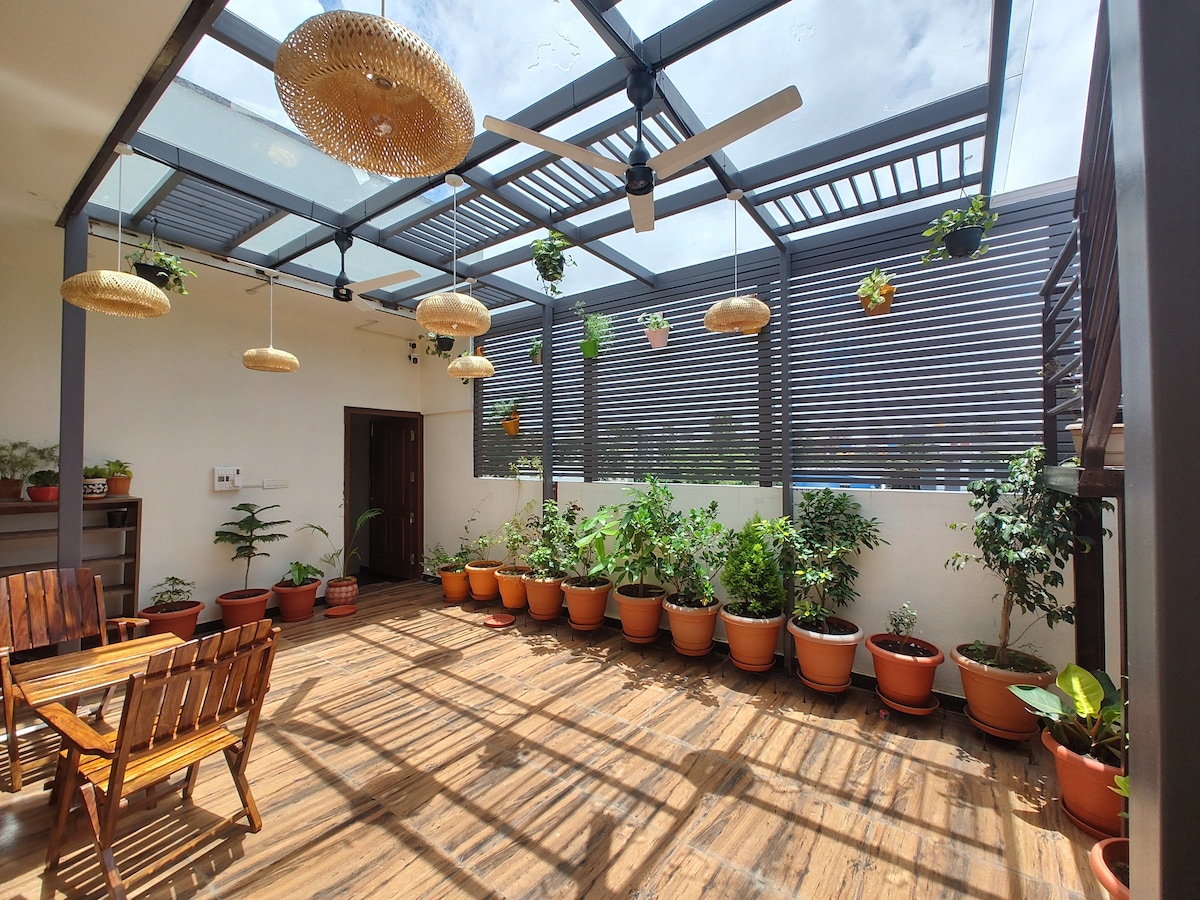 A spacious terrace is adorned with numerous potted plants, adding greenery to the inviting atmosphere. Wooden seating is arranged for outdoor relaxation. The area is illuminated by natural light filtering through a glass ceiling, complemented by decorative ceiling fans and hanging light fixtures.