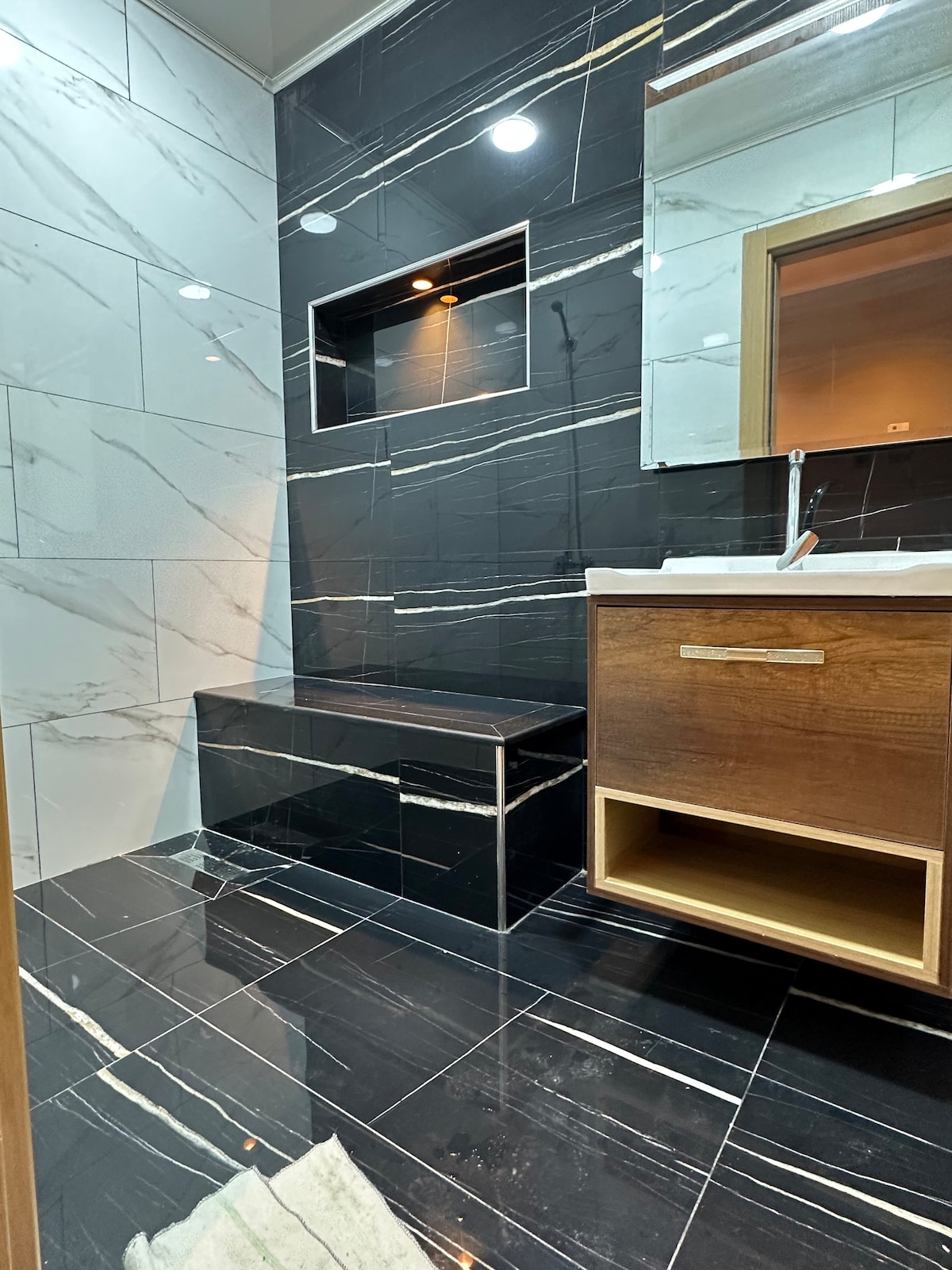 The bathroom features sleek black and white marble tiles that create a modern aesthetic. A wooden vanity with an open shelf is positioned under a mirror, while a built-in recess provides a space for toiletries. Soft lighting enhances the overall design.