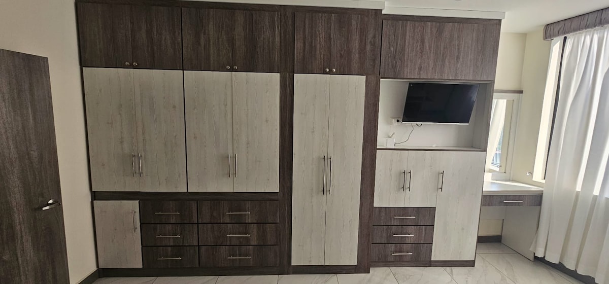 A wall unit composed of cabinetry in light and dark wood tones occupies a portion of the room. Several drawers and cupboards provide ample storage space. A flat-screen television and a mirror are integrated into the design, with natural light flowing in from a nearby window covered by white curtains.
