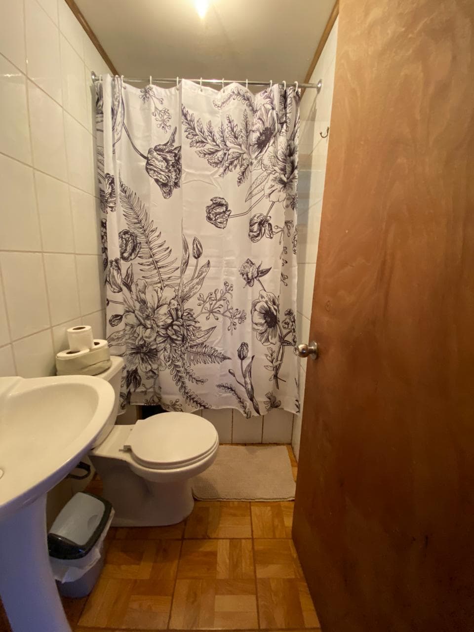 A clean bathroom features a white sink and a toilet. A floral shower curtain adds a decorative touch, while neutral tiles and a wooden door complete the space. Towels and toilet paper are neatly arranged, ensuring convenience for guests.