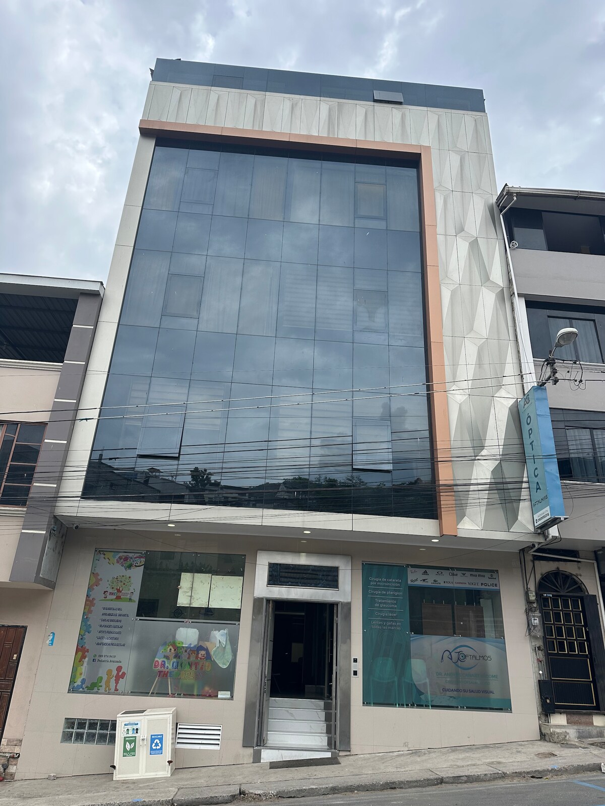 A modern building with a sleek glass façade features a prominent geometric design. Large windows allow natural light to fill the interior, while ground-level displays showcase colorful signage. The entrance is marked by a minimalist door and a hint of decorative elements.