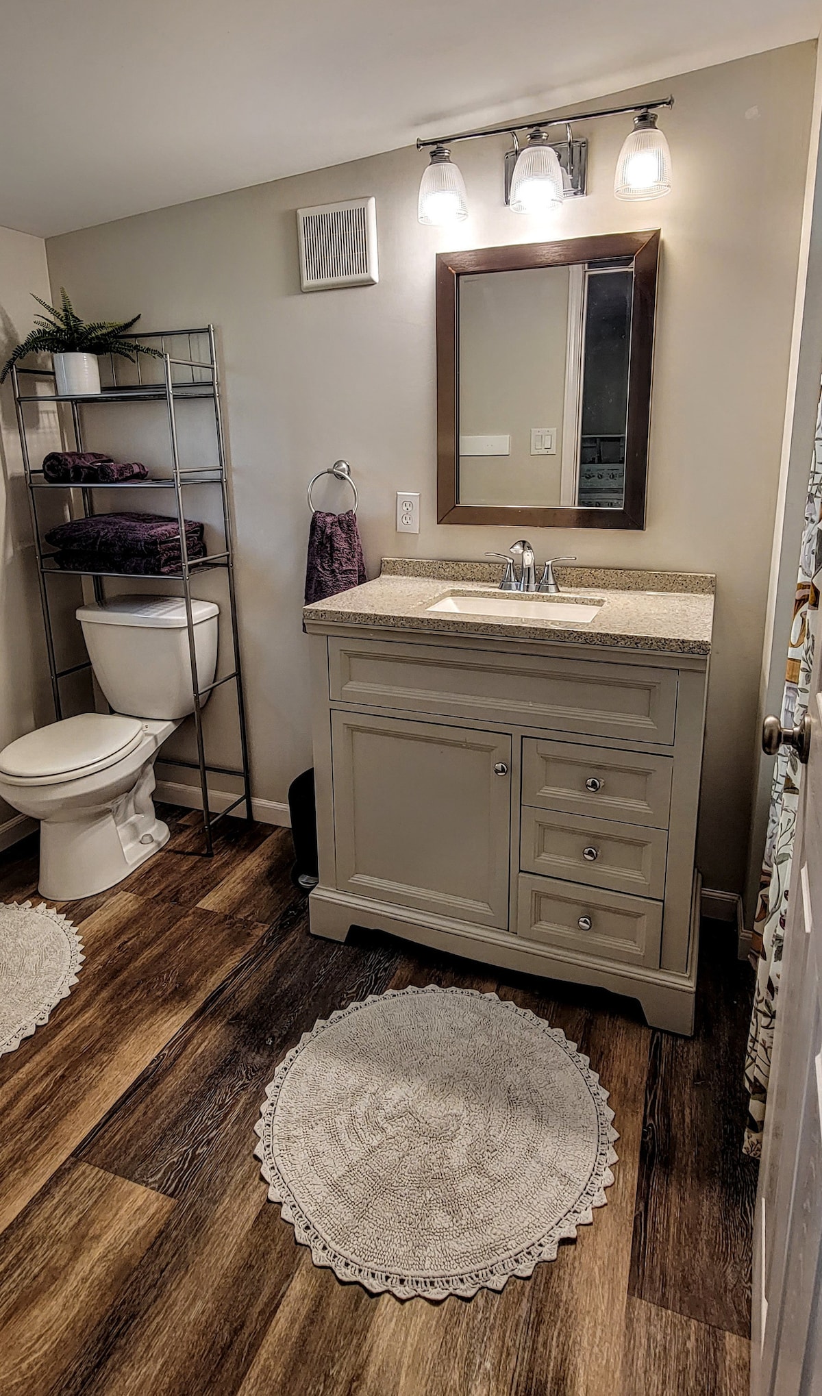 A clean bathroom features a modern vanity with a granite countertop and a large mirror above. Shelving holds neatly stacked towels. A toilet is positioned adjacent to the vanity. The floor is covered with round, textured rugs, complementing the overall design.