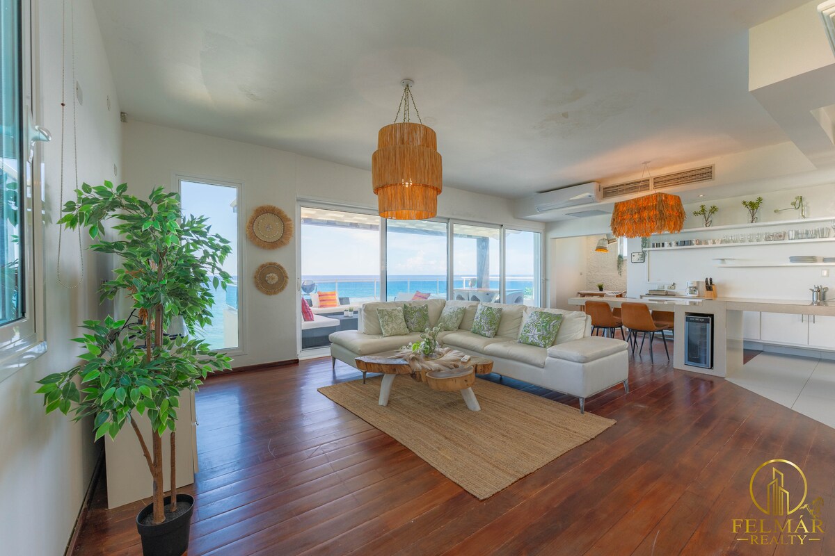 A spacious living area features large windows that open to ocean views. A light-colored sofa is positioned around a wooden coffee table, with decorative pillows enhancing the seating area. Natural light fills the space, complemented by stylish light fixtures and indoor greenery.
