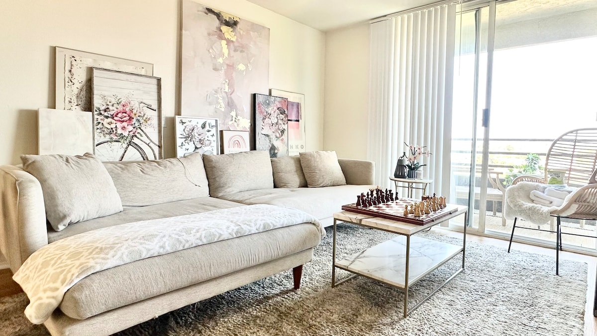 A light-filled living area features a comfortable sectional sofa draped with a soft blanket. A glass-top coffee table is positioned atop a plush rug, showcasing a chess set. Framed artwork adorns the walls, and sliding glass doors lead to a balcony with an outdoor chair.