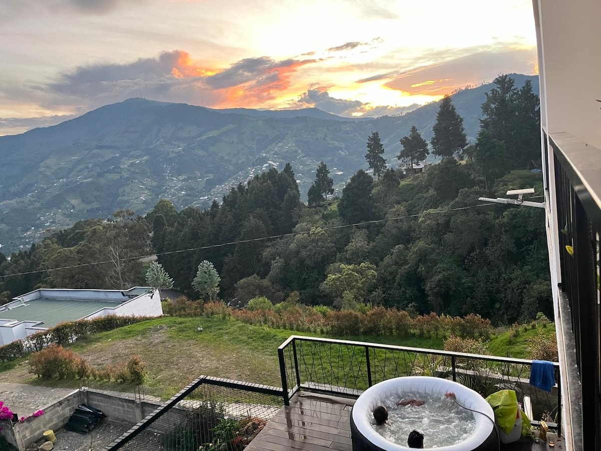 A scenic outdoor terrace features a hot tub overlooking a mountainous landscape. The sky displays a range of colors as the sun sets, reflecting the tranquil atmosphere. Lush greenery surrounds the area, providing a peaceful backdrop for relaxation.