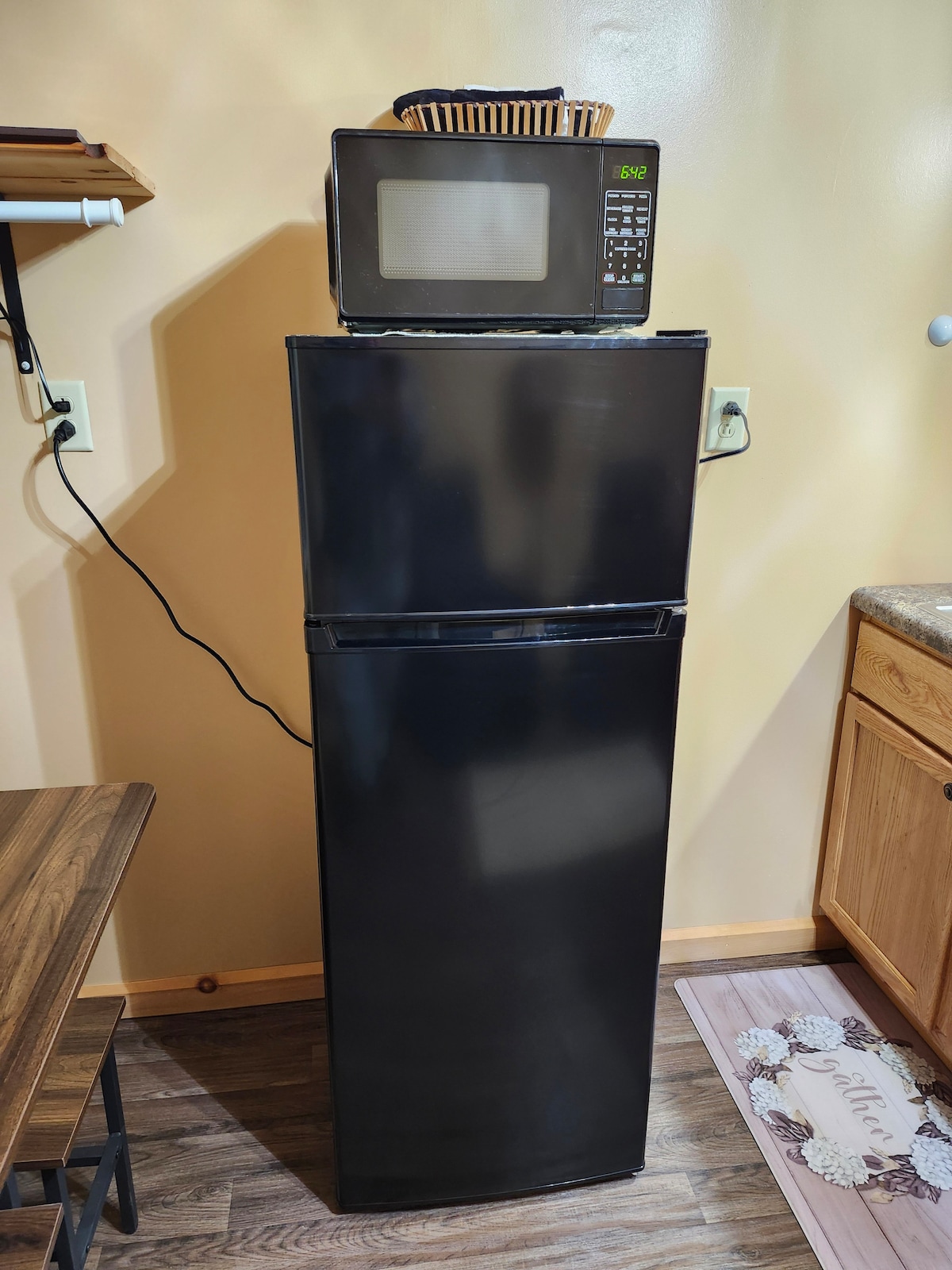 A black refrigerator with a microwave placed on top is positioned against a light-colored wall. Next to it, a small countertop is visible with wooden cabinets below. A kitchen mat is partially seen on the floor, complementing the overall kitchen setup.