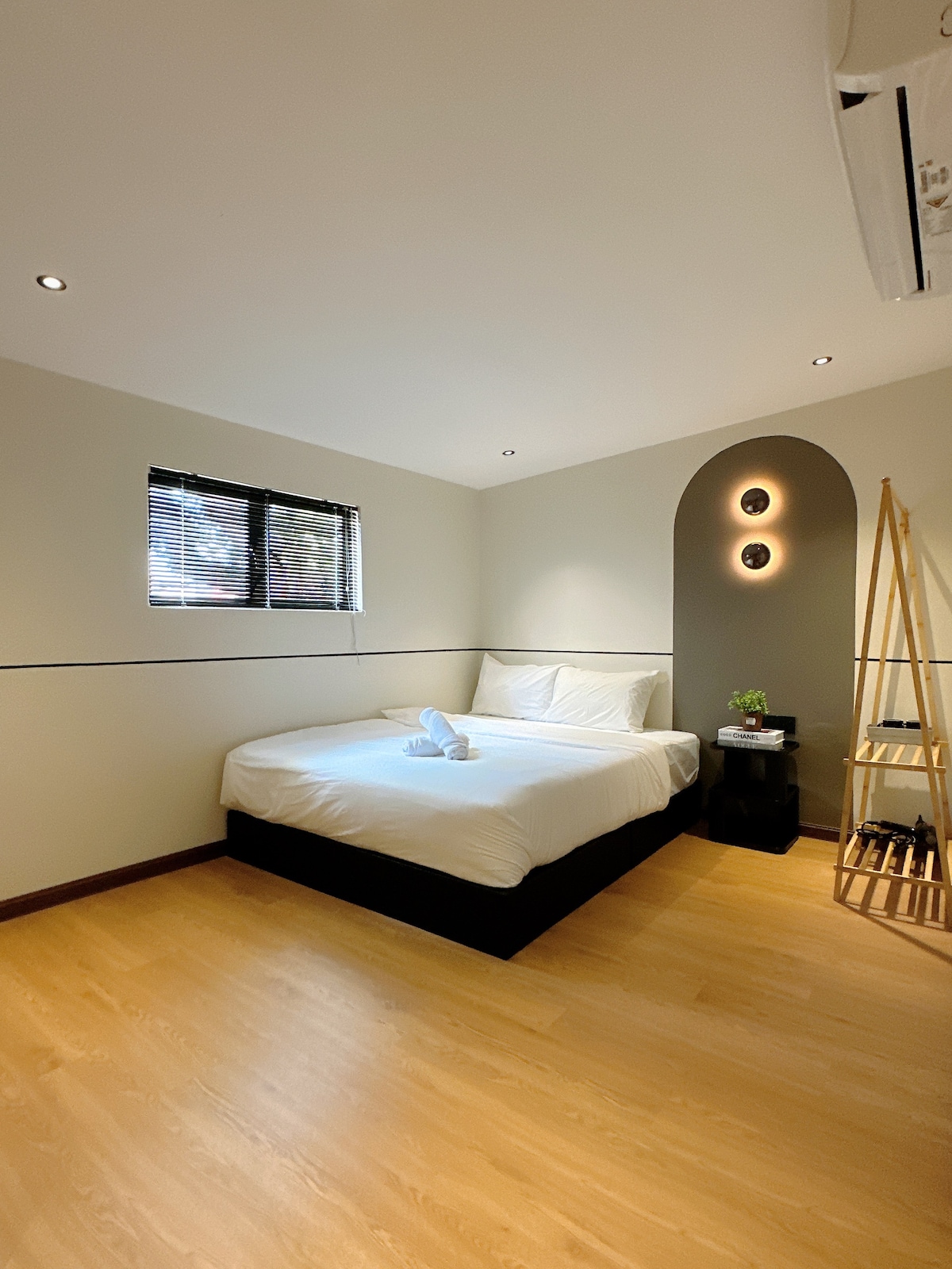 A private room features a spacious bed, complemented by soft linens. Natural light filters through the window, covered with blinds. A stylish shelving unit stands against the wall, and decorative accents include two wall lights, enhancing the room's ambiance.