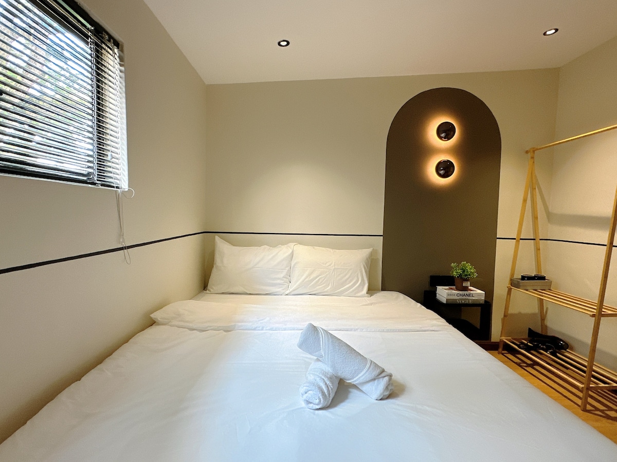 The private room features a spacious bed with crisp white linens and neatly rolled towels. Natural light enters through a window fitted with blinds, enhancing the simple design, which includes a minimalistic wooden shelf and unique wall lighting.
