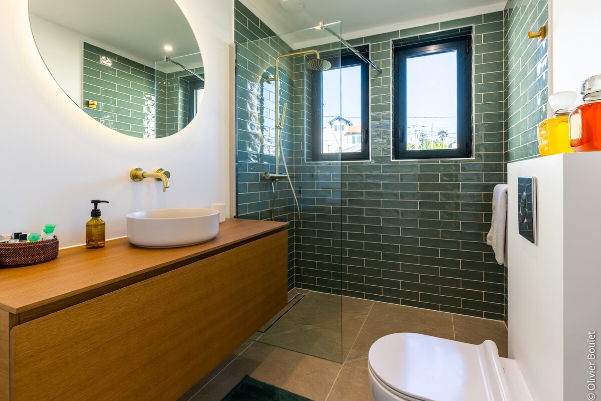 The bathroom features a modern design with green tile walls and a sleek wooden vanity. A round mirror is mounted above the basin, while a walk-in shower is equipped with a rain showerhead. Natural light enters through two windows, enhancing the spacious feel.