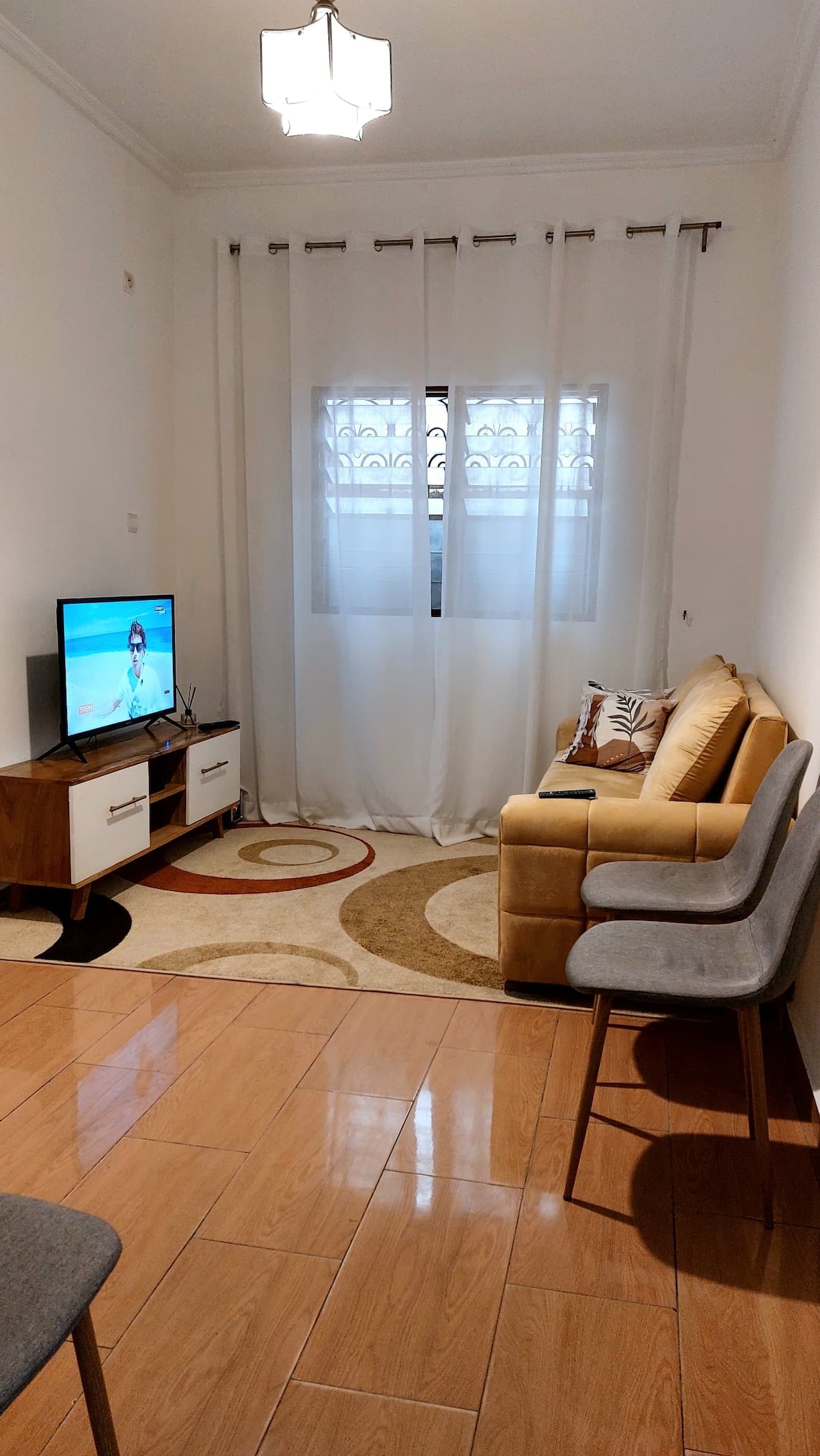 A bright living area features a cozy seating arrangement with a beige sofa and two grey chairs. A wooden TV stand holds a television, while a circular rug adds warmth to the space. Natural light filters through sheer curtains covering a window.