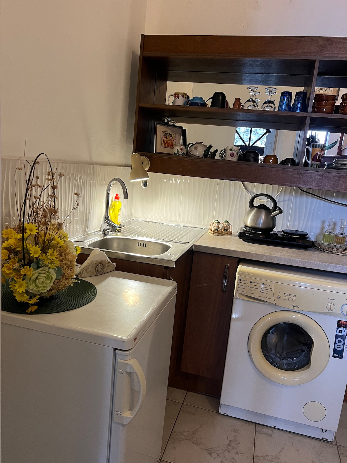 A compact kitchen area features a blend of modern and rustic elements. A stainless steel sink is placed next to a white refrigerator and a washing machine. Shelves display cups and decor, while a kettle sits ready on the stovetop. Warm lighting highlights the functional design.