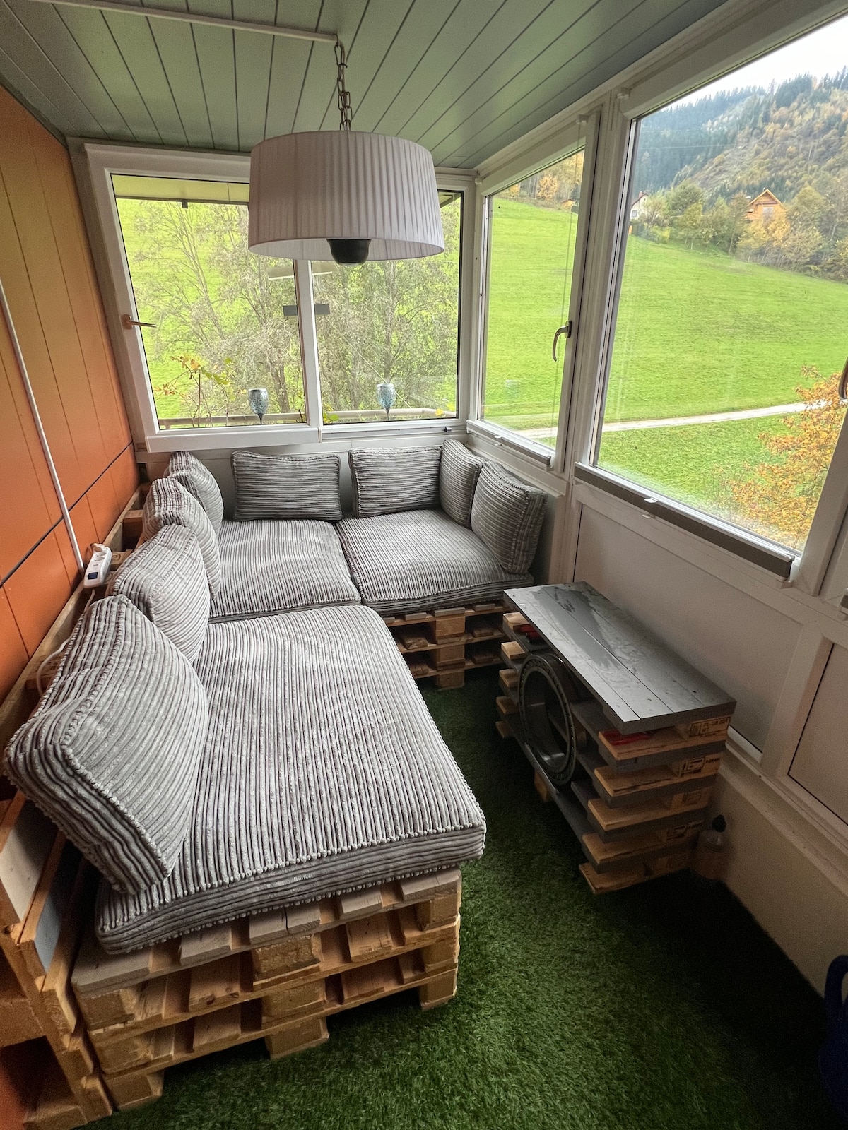 A sunlit seating area is enclosed by windows, featuring a cozy corner sofa crafted from pallet furniture, adorned with striped cushions. A small grass-like rug covers the floor, and a pendant lamp hangs overhead, creating a relaxed space with views of the green landscape outside.