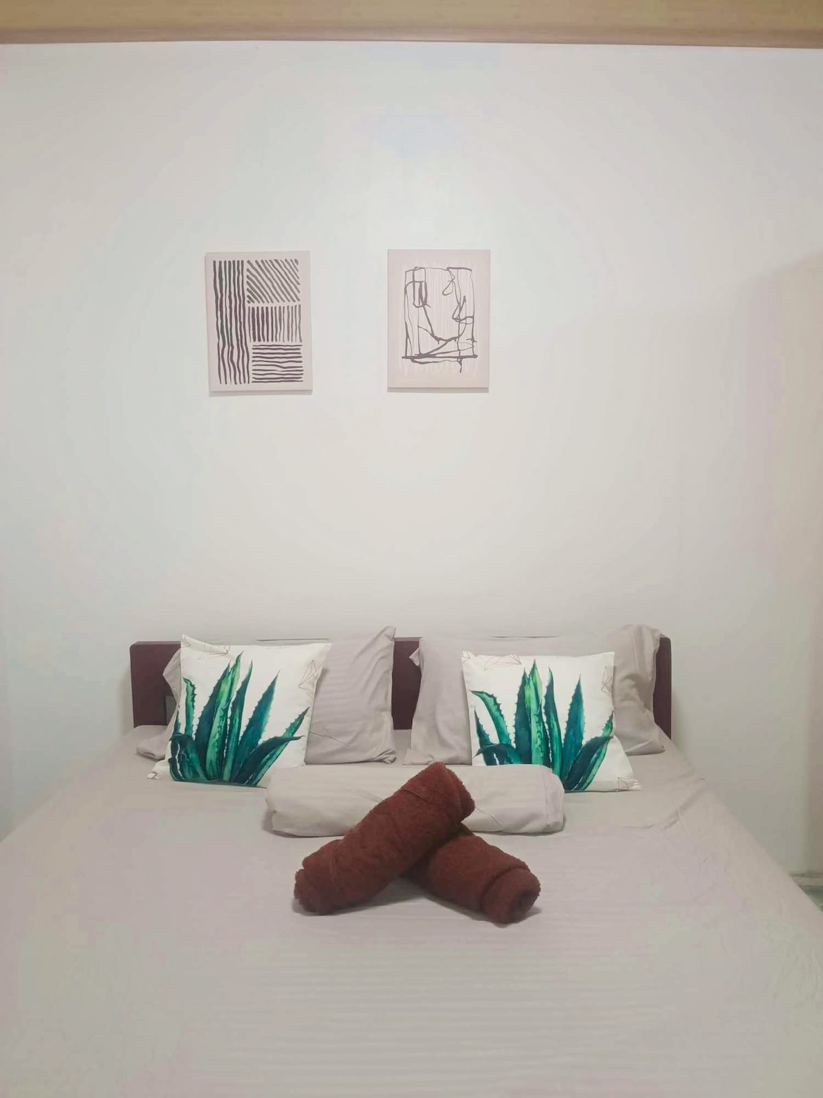 The bedroom features a spacious bed adorned with decorative pillows and rolled towels. Two framed artworks are displayed on the white wall above the bed, complementing the room's simple design.