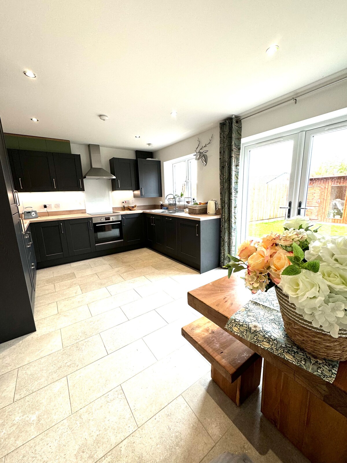 A modern kitchen features dark cabinetry and a light countertop, creating a stylish contrast. A wooden dining table is complemented by a vase filled with soft-colored flowers, inviting a casual dining experience. Natural light streams in through sliding glass doors, illuminating the space.