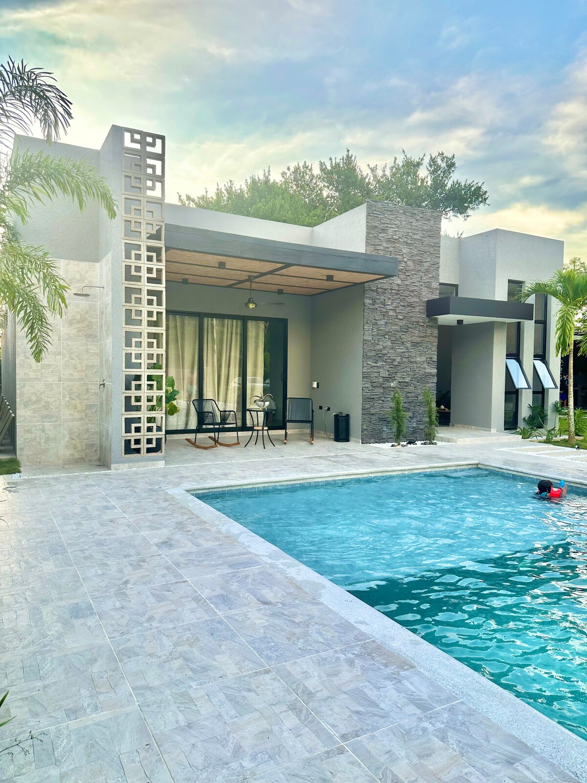 A serene outdoor space is featured, with a clear blue swimming pool reflecting the soft sunlight. A cozy seating area is positioned under a covered patio, adjacent to the pool, with a stone accent wall adding character to the modern exterior.