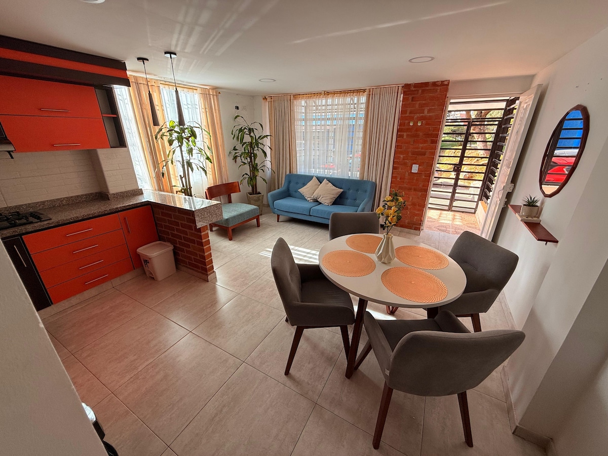 A modern kitchen and dining area is visible, featuring a round table set for four. Bright orange cabinets contrast with the neutral tones of the tiled floor. Large windows allow natural light to fill the space, accompanied by a cozy blue sofa and decorative plants.