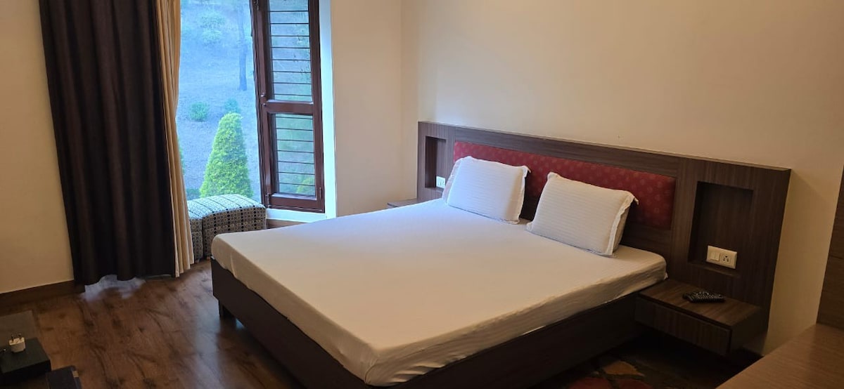 A spacious sleeping area features a neutral-colored bed with crisp white linens. Natural light filters in through a window adorned with wooden shutters, illuminating the wooden flooring. A small seating area is visible beside the window, adding to the room's serene ambiance.