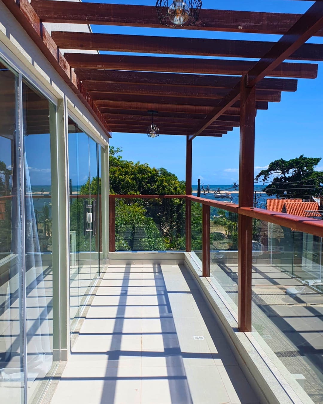 A well-lit balcony features wooden beams overhead, providing shade and a view of the surrounding trees and ocean. Glass railings create an open feel, and sunlight casts shadows across the tiled floor, enhancing the spacious ambiance.