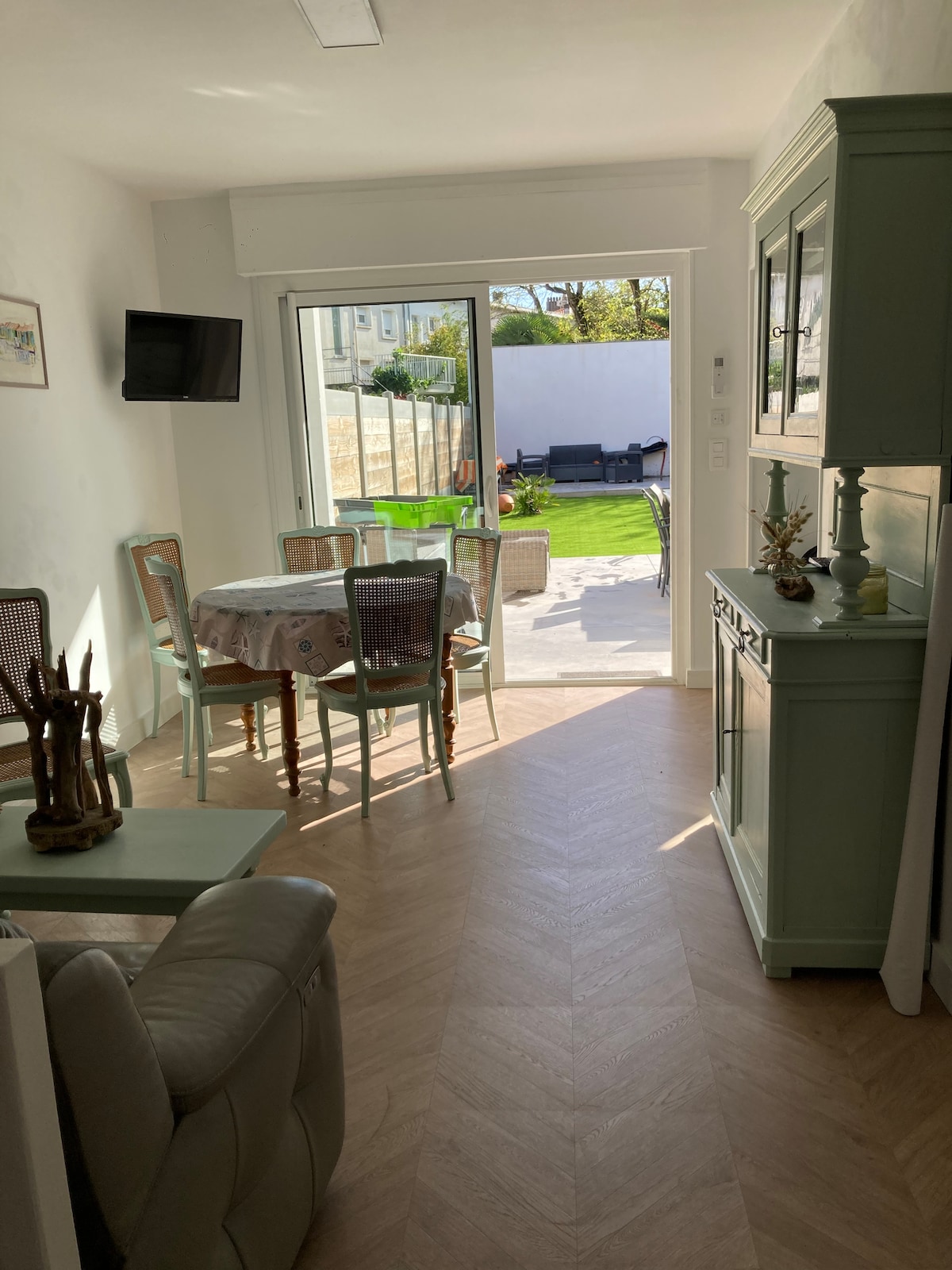 A bright dining area features a round table surrounded by four chairs, with large glass doors opening to a landscaped patio. A light green cabinet adds a soft touch, while a comfortable seating area is visible in the corner, enhancing the welcoming space.