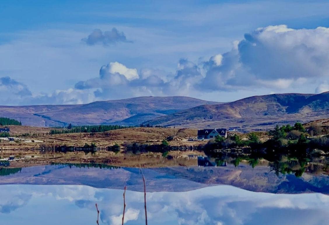 A tranquil lake reflects the surrounding hills and sky, creating a serene landscape. Rolling mountains line the horizon, with patches of greenery dotting the lower slopes. A few distant buildings are visible, subtly integrated into the natural scenery.