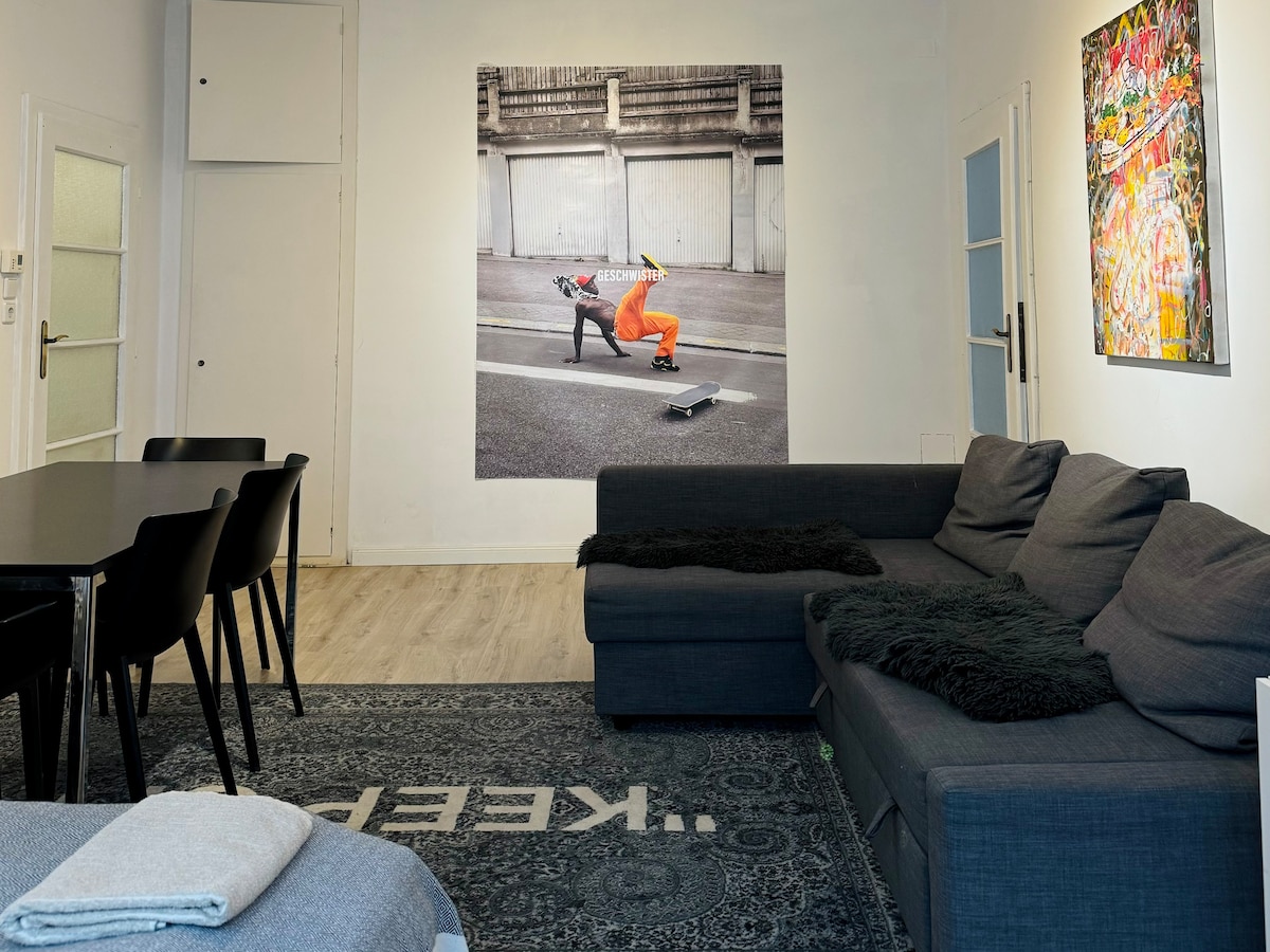 A modern living area features a large gray sectional sofa and a black dining table. Artistic wall murals, including a skateboarder, enhance the space. A patterned area rug lies beneath, contributing to the unique decor, while light colors create an open and inviting ambiance.