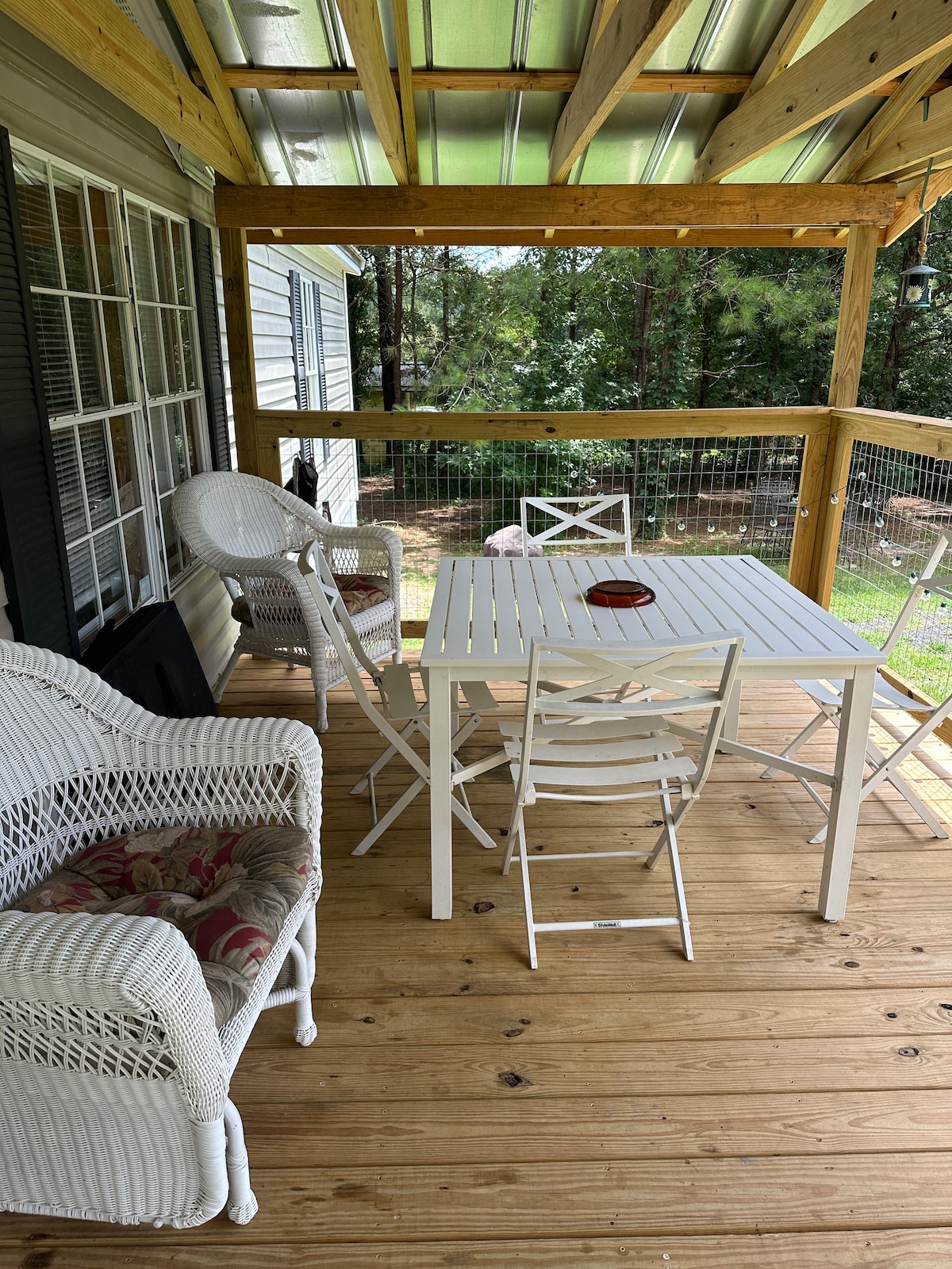 A shaded porch features a white table surrounded by four folding chairs. Two wicker chairs are positioned nearby, inviting relaxation. Natural wood flooring adds warmth, while greenery from the yard is visible through the railing. The space is designed for casual gatherings and conversation.