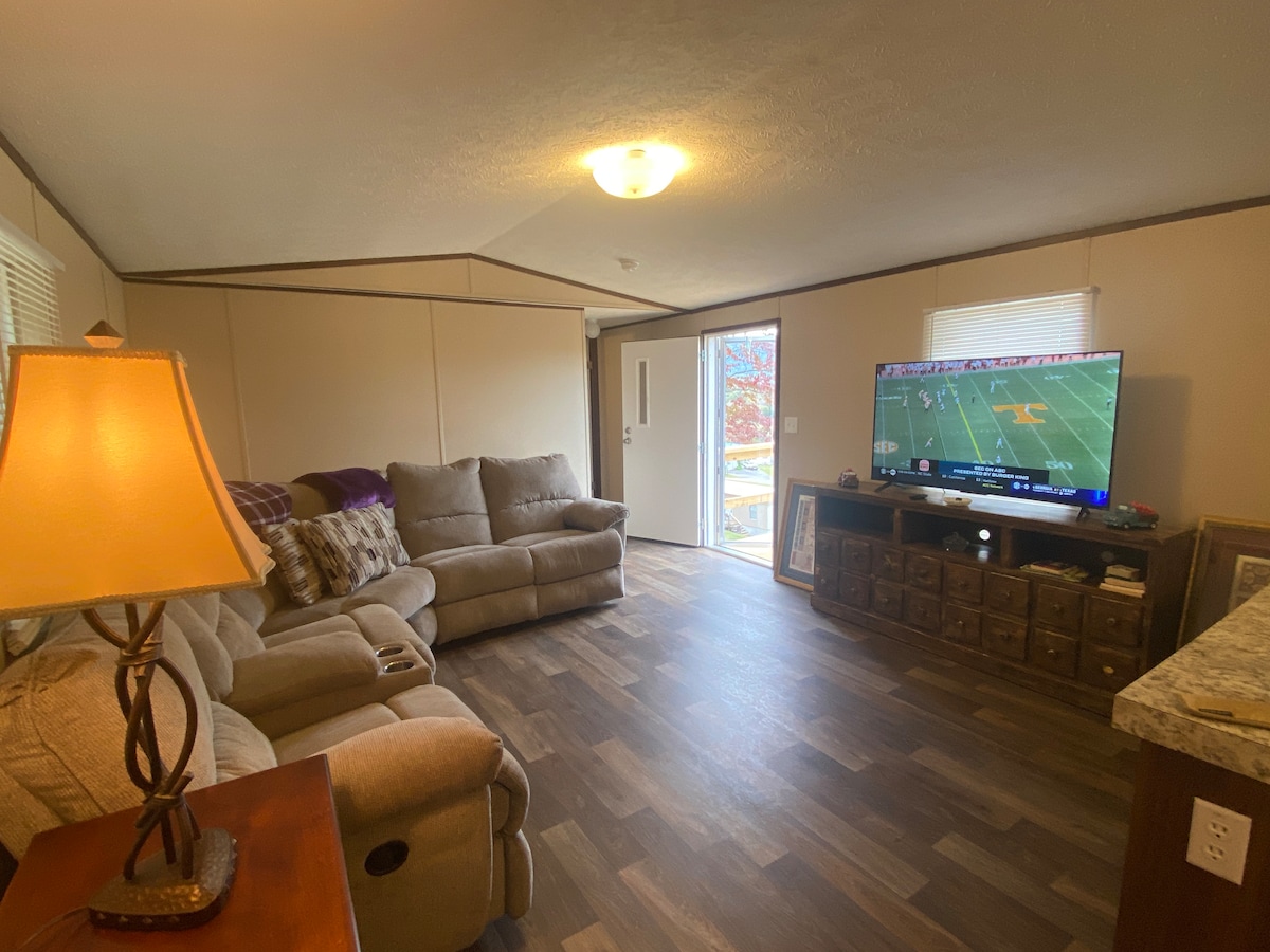 A spacious living area is furnished with a comfortable sectional sofa and a coffee table. A flat-screen TV displays a sports broadcast. A door leads to the outdoors, while natural light filters through windows on either side, illuminating the warm tones of the decor.