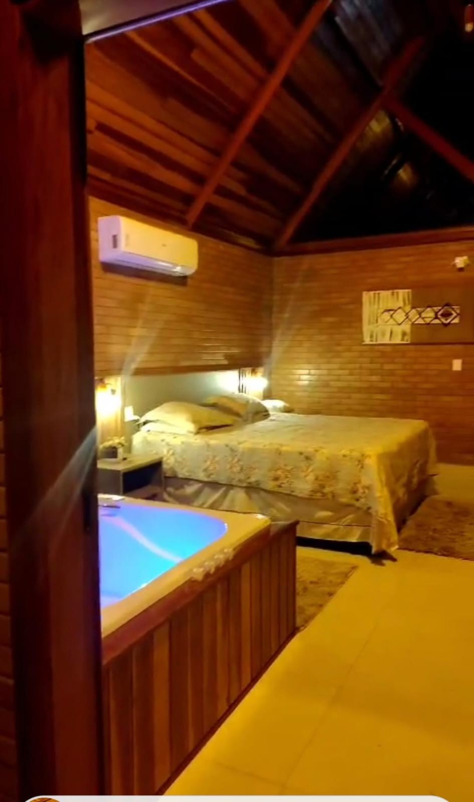 A cozy room is presented with a spacious bed adorned in floral bedding. A softly illuminated hot tub is positioned nearby, creating a relaxing atmosphere. Wooden beams and brick walls add warmth, while an air conditioning unit is mounted above for comfort.