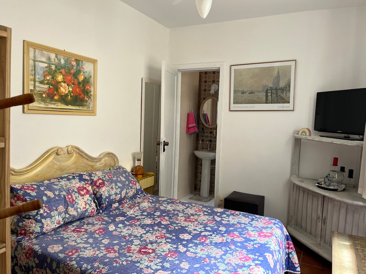 A bright bedroom features a spacious bed adorned with a floral-patterned blanket. A wooden dresser and a stable surface are present. A door leads to an adjoining bathroom, and a small television is positioned on a white entertainment unit against the wall.