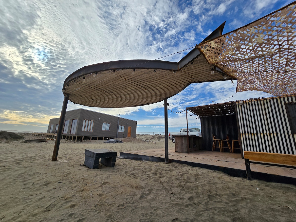 A shaded outdoor area is depicted with a curved wooden roof supported by pillars. Sand covers the ground, and a seating area is visible with chairs and a table. A simple structure can be seen in the background, surrounded by the beach landscape.