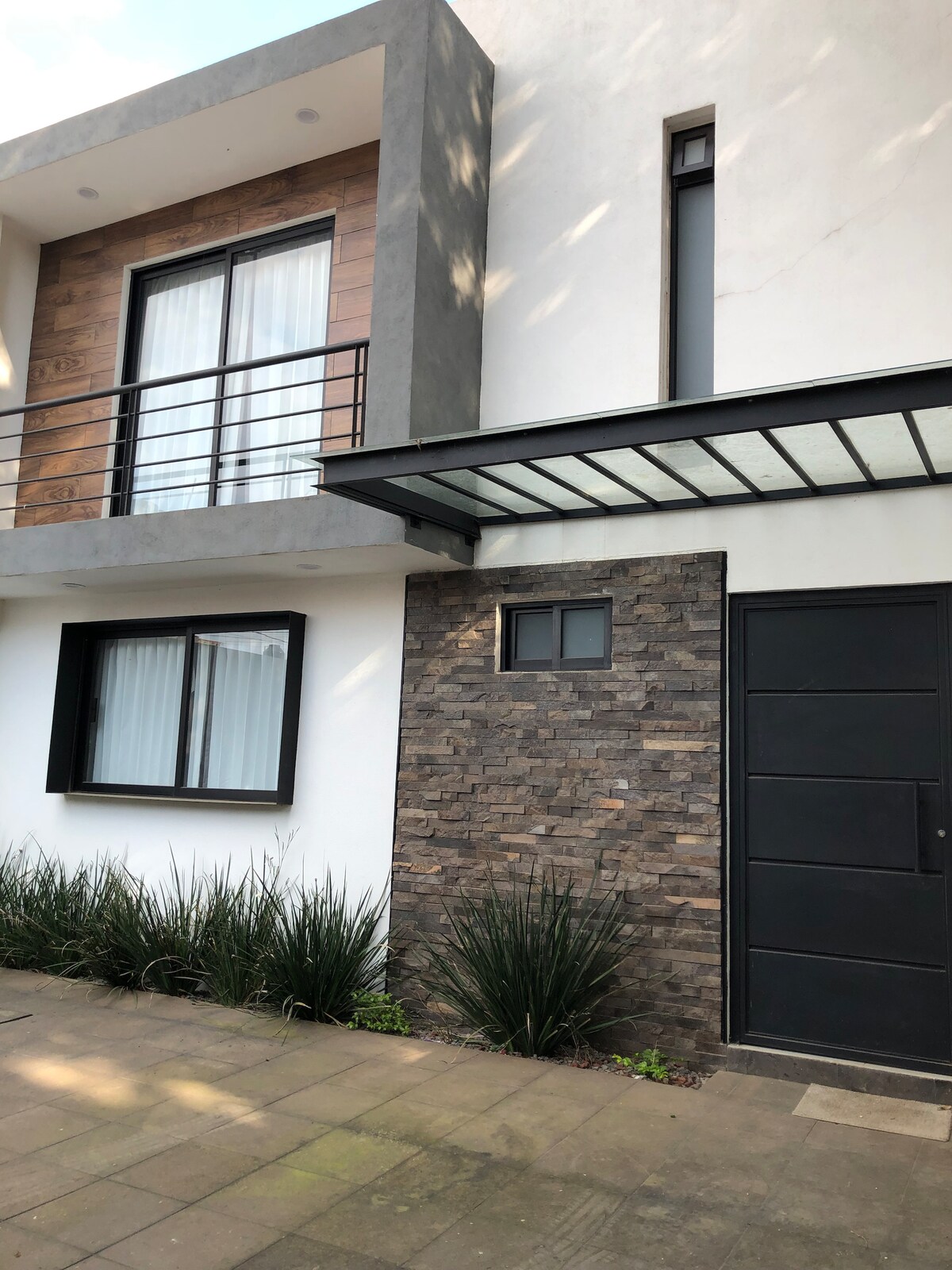 A modern building façade is presented, featuring a combination of textured stone and smooth white walls. Large windows provide natural light, with a balcony extending from the upper level. Minimalistic landscaping with low greenery enhances the entrance area.