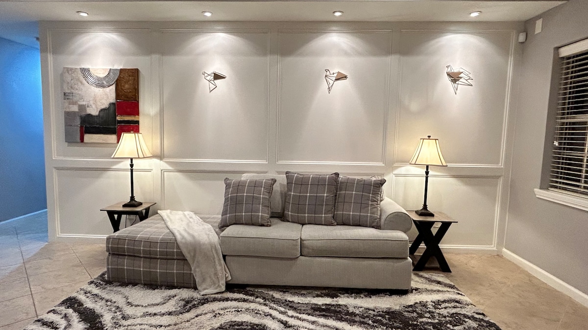 A spacious living area is presented, featuring a light gray sectional sofa adorned with decorative pillows. Two elegant table lamps flank the sofa, casting a soft glow. The textured area rug enhances the space, while artwork on the wall adds visual interest.