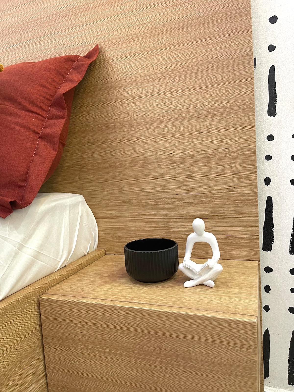 A bedside table is positioned next to a spacious bed, featuring a small black bowl and a white decorative figure sitting serenely. The headboard is made of light wood, complemented by warm red cushions, creating a calming and minimalist design.