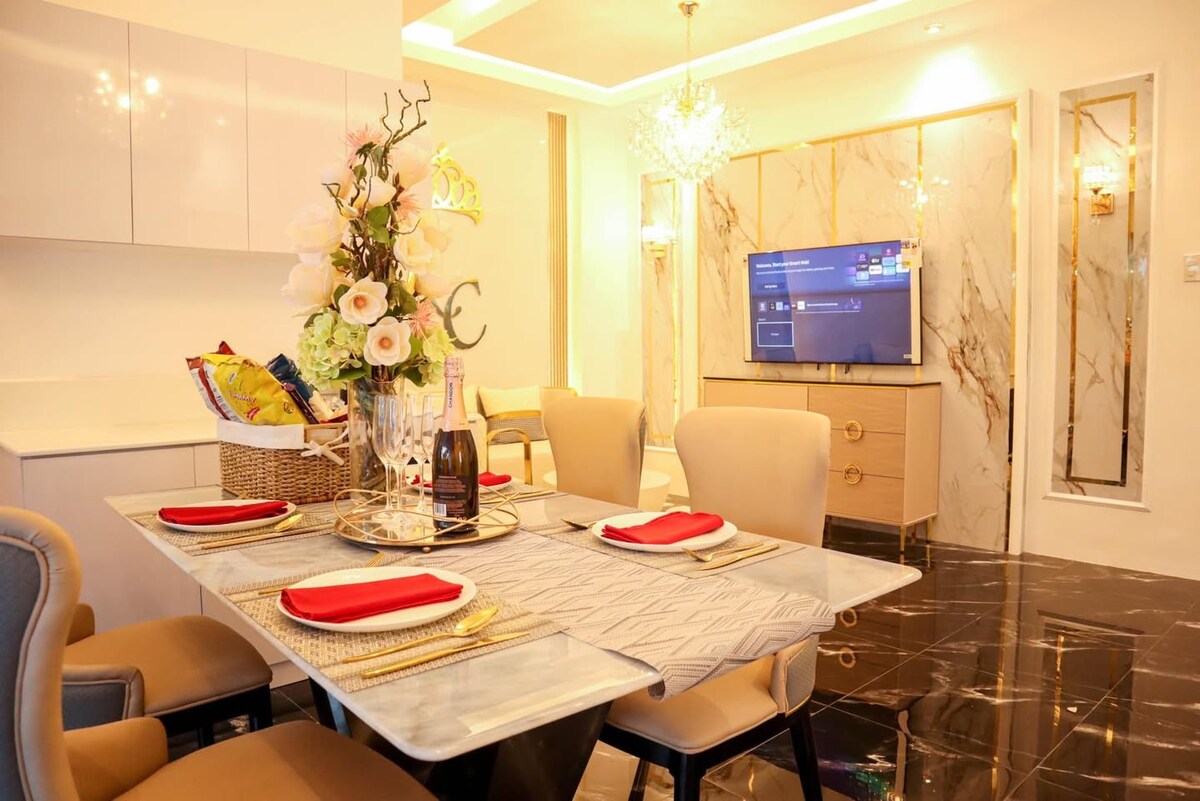 A stylish dining area features a glass-top table set for four, with elegant tableware and flower arrangements. Soft lighting is provided by a chandelier and wall sconces, while a television is mounted on a tastefully designed feature wall in the background.