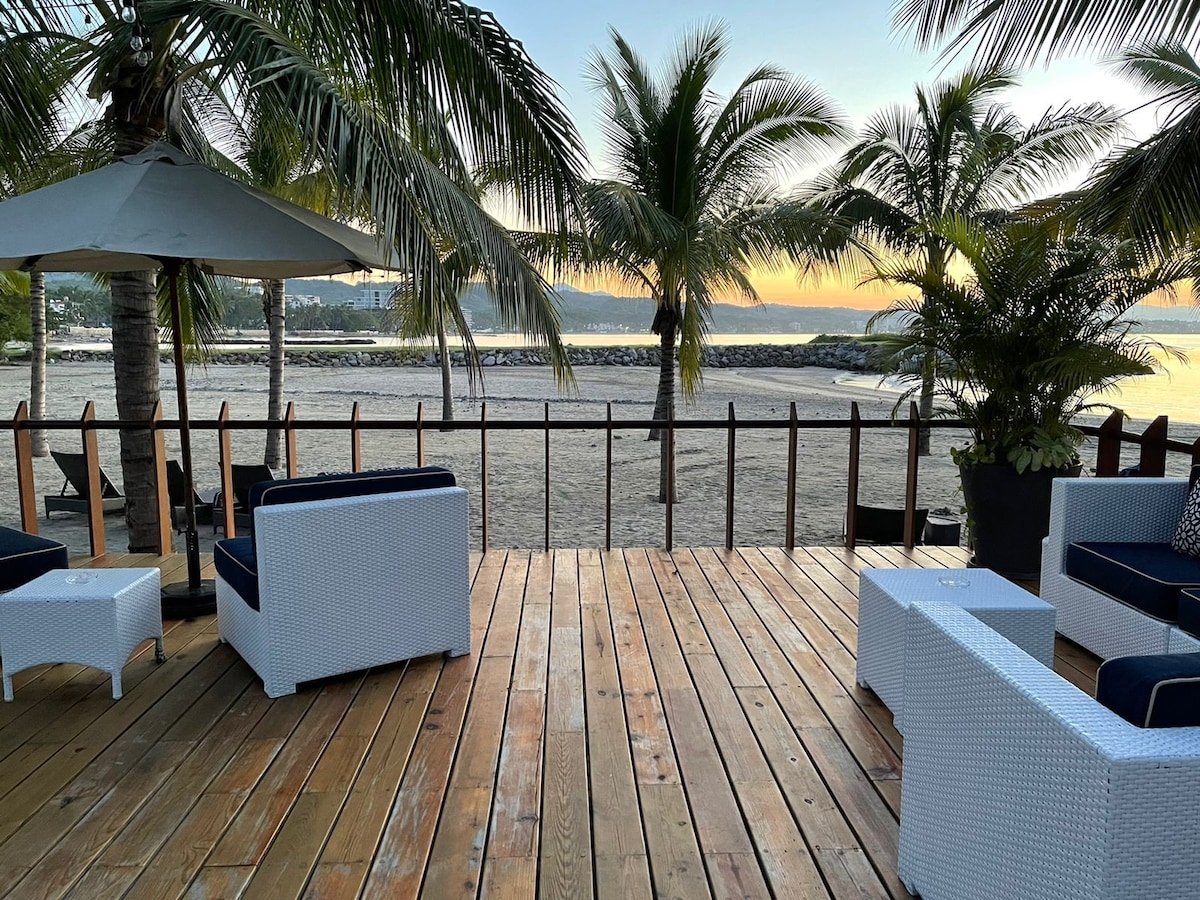 An outdoor lounge area is presented, featuring white wicker furniture arranged on a wooden deck. Palm trees gently sway in the breeze, framing the view of a sandy beach and tranquil water. The early morning light casts a soft glow over the scene.