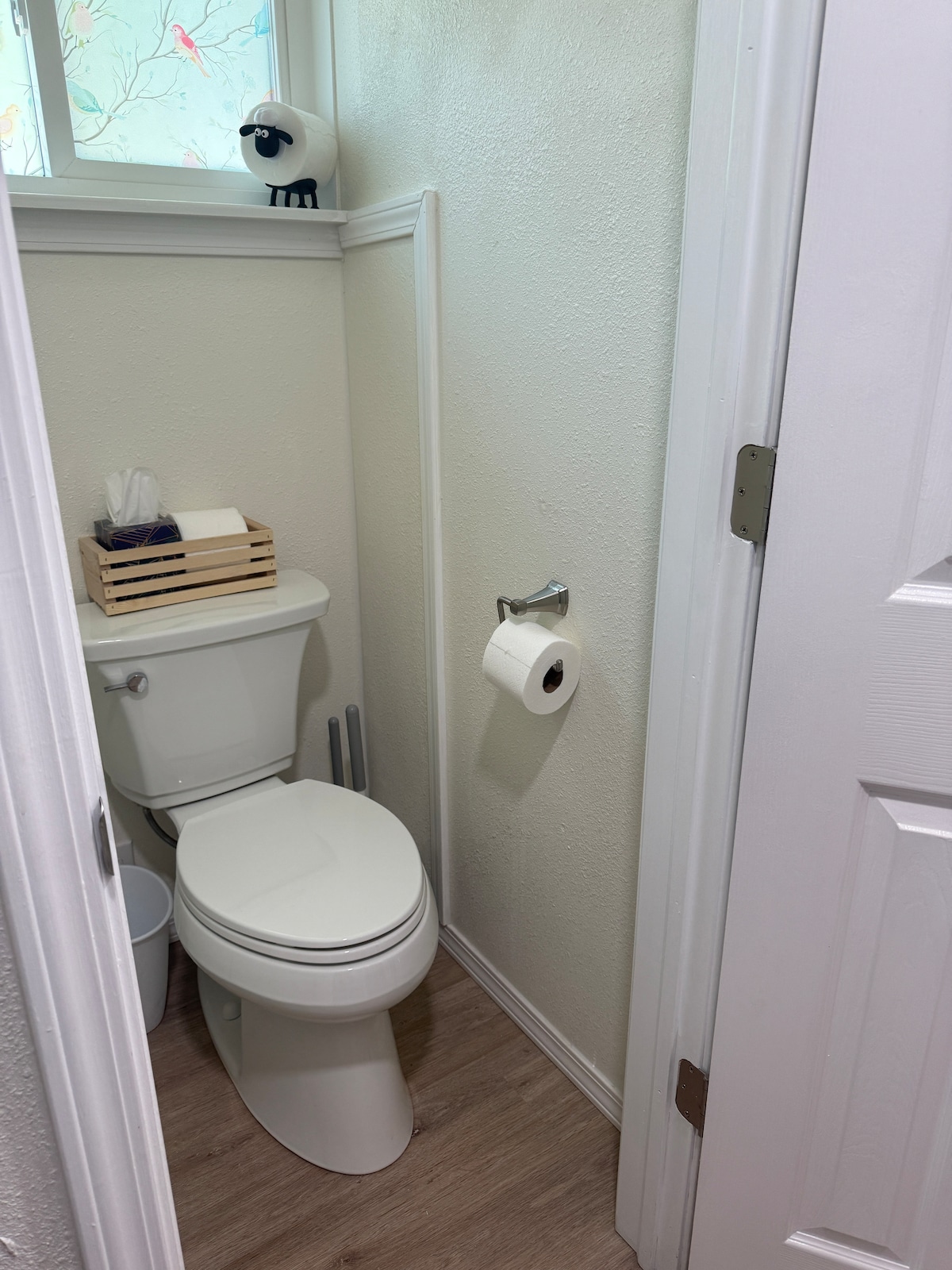 A compact bathroom space is equipped with a standard toilet and a small wastebasket. A wooden tray holds tissue and personal care items, while a roll of toilet paper is mounted on the nearby wall. Natural light enters through a partially frosted window.