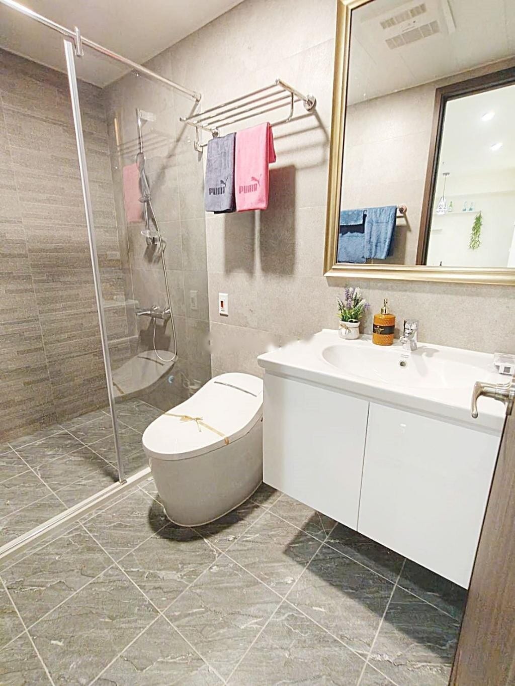 A modern bathroom features a glass shower enclosure with gray tile accents. A white vanity with a sink is positioned beside a minimalistic toilet. Towel racks display neatly organized towels in various colors. Natural light enters through a nearby window, enhancing the clean design.