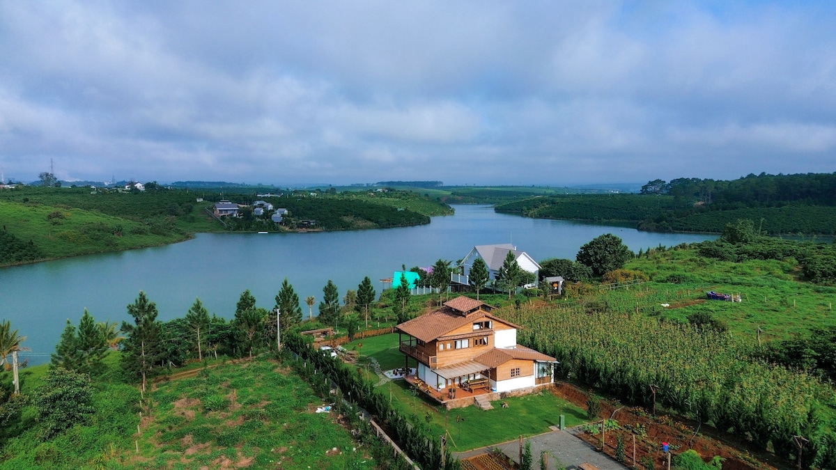 A lakeside property is visible with a wooden house surrounded by greenery and water. Lush fields and trees border the tranquil lake, where the sky reflects a soft blue hue. Nearby, a cornfield can be seen, contributing to the serene rural landscape.