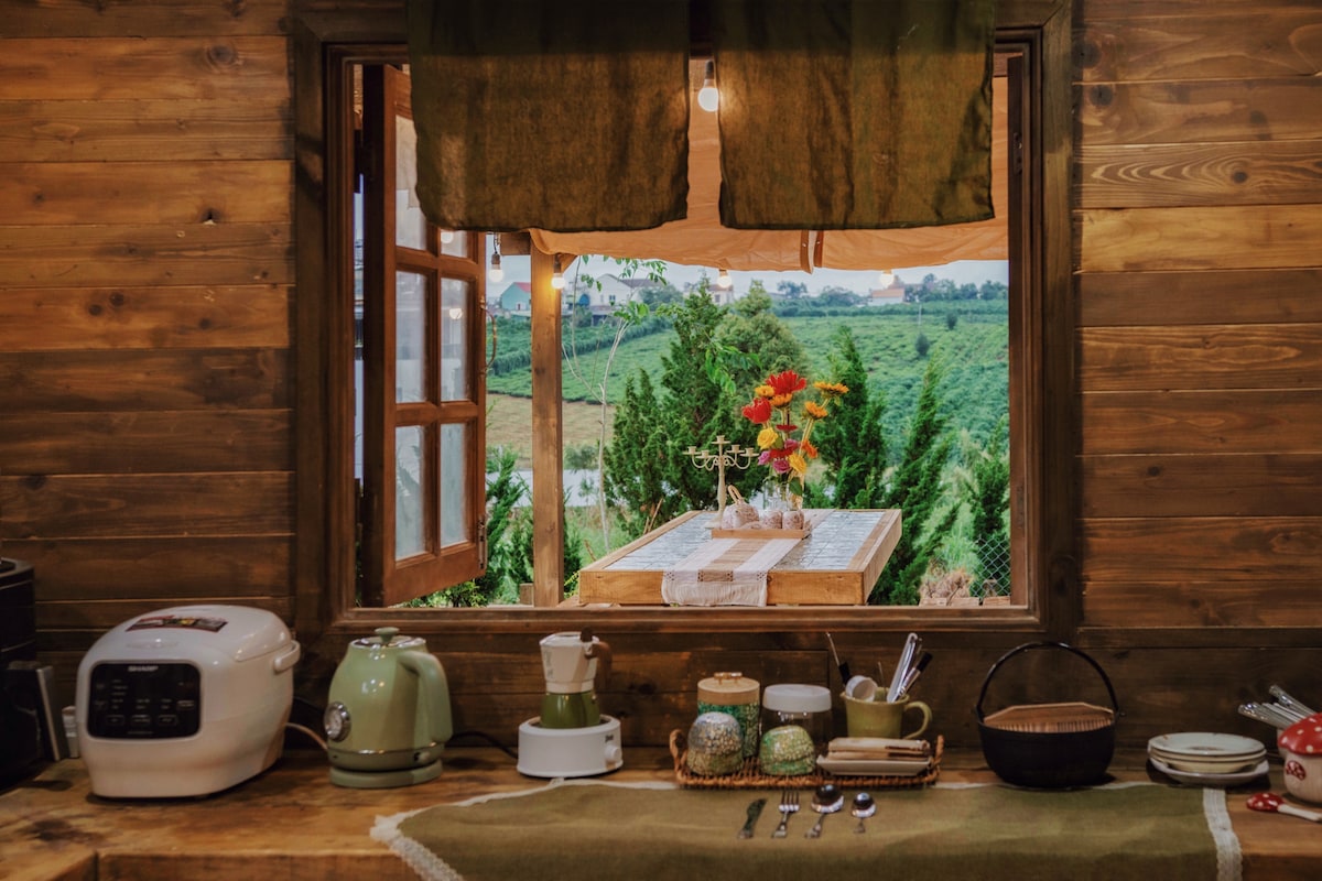 A rustic kitchen window reveals a view of green hills and a dining setup outside. The space includes a variety of kitchen appliances, utensils, and decorative elements. Soft light filters through the curtain, enhancing the inviting atmosphere of the room.