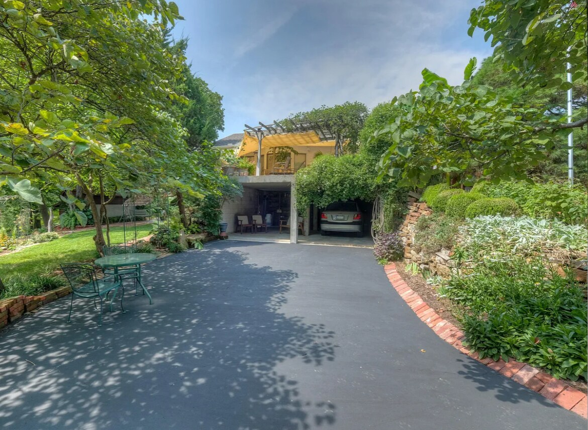 A welcoming entrance features a spacious driveway bordered by lush greenery and garden beds. A covered area provides shelter for vehicles, while a seating arrangement is positioned on the right, inviting relaxation in a serene outdoor setting.