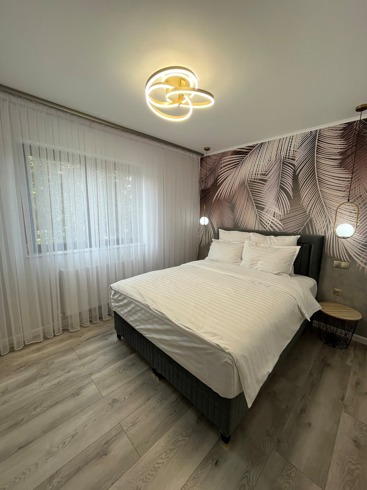 A well-lit bedroom features a spacious bed with white linens and a leafy wallpaper accent. Soft light is provided by a modern ceiling fixture, while sheer curtains frame the window, allowing natural light to filter in.
