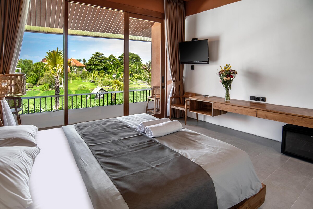 A spacious bedroom features a large bed with a neutral-toned blanket, prominently displayed against a scenic backdrop. Floor-to-ceiling windows frame lush greenery outside, while a flat-screen TV is mounted on the wall. A wooden console holds a small bouquet of fresh flowers.
