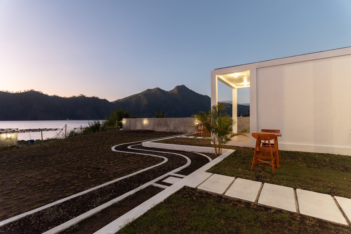 An outdoor space features a well-kept lawn bordered by a white pathway leading to a small wooden table. A serene landscape of mountains is visible in the background, with soft evening light enhancing the natural surroundings.