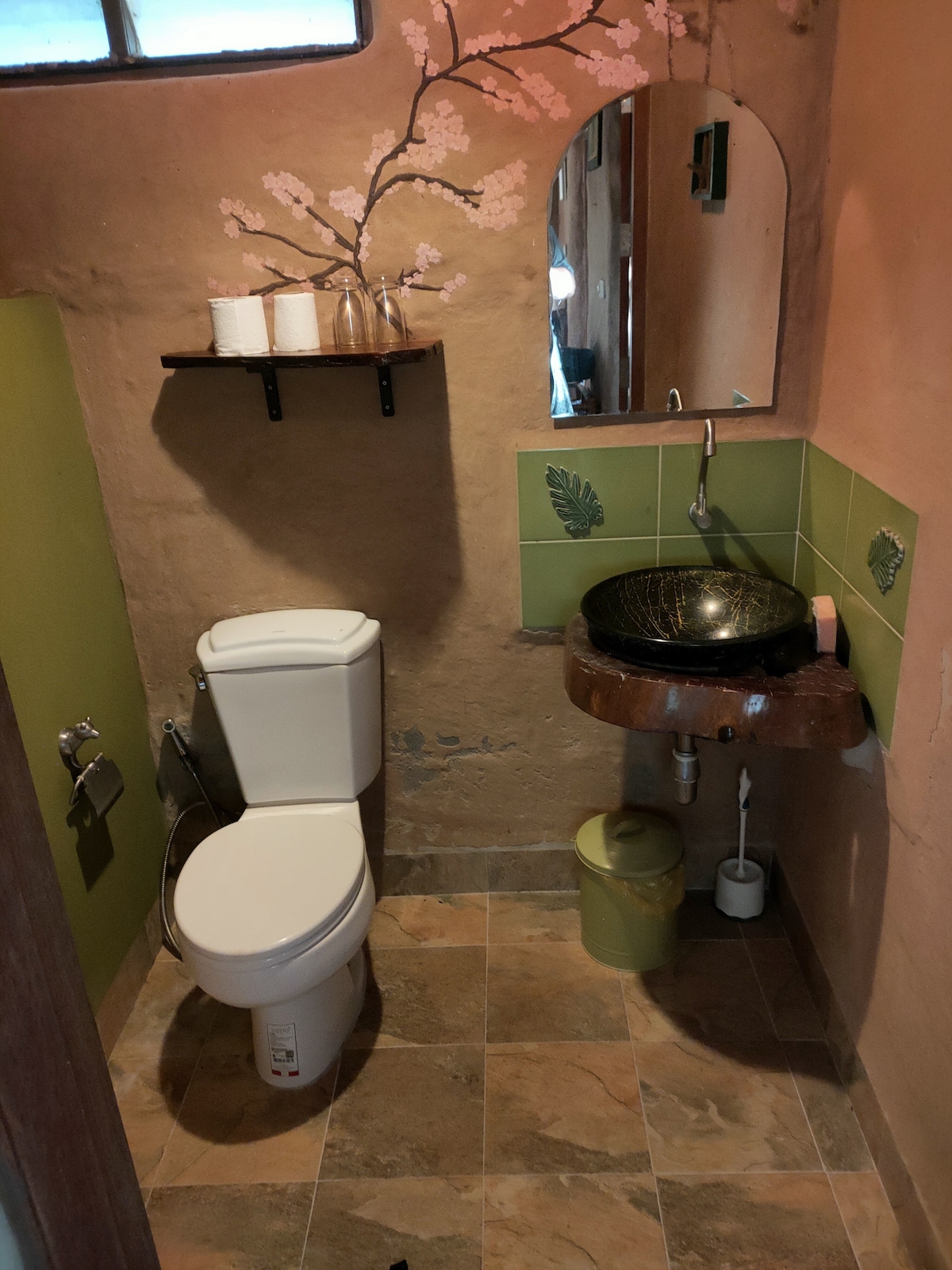 A cozy bathroom space features earthy tones with a textured brown wall and green accents. A circular, stone sink rests on a wooden countertop, while a mirror above reflects the gentle light. A small shelf holds toiletries, and a modern toilet is positioned adjacent.