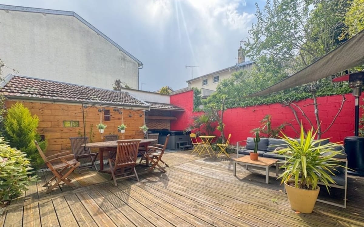 A spacious terrace features wooden decking and is surrounded by greenery. A wooden table with matching chairs is positioned at the center, while casual seating areas are arranged nearby. A vibrant red wall adds a modern touch, complemented by potted plants and outdoor heating.