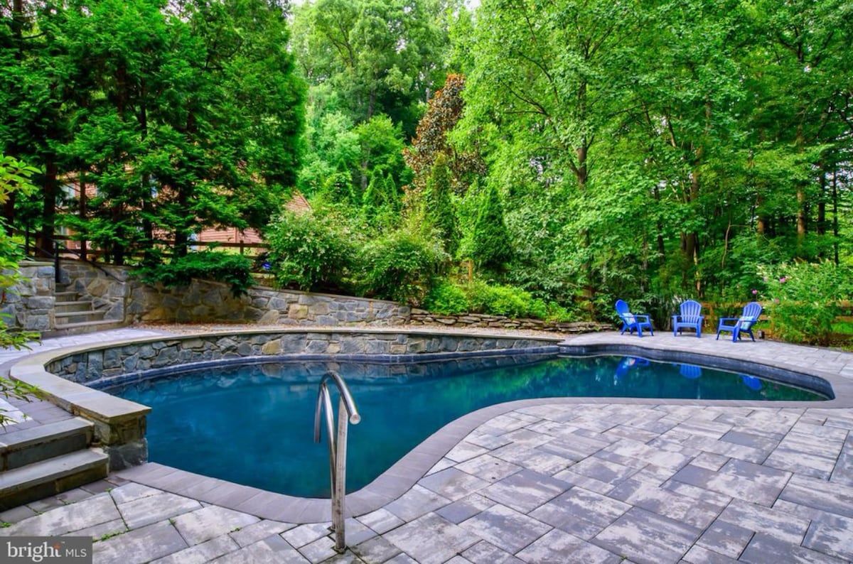 A serene backyard features a gently curved pool, surrounded by lush greenery and mature trees. Blue adirondack chairs are positioned on the stone patio, inviting relaxation amid the tranquil natural setting. The pool's water reflects the vibrant colors of the surrounding landscape.