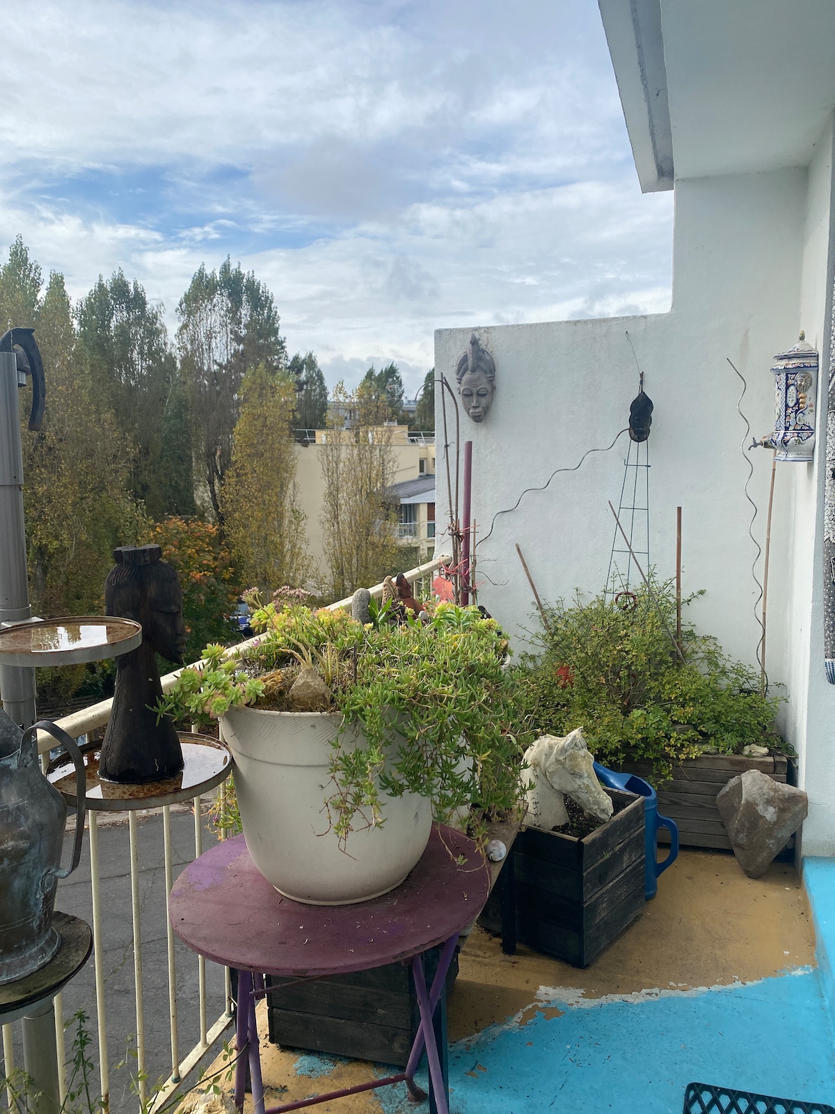 A balcony space is adorned with various potted plants, creating a lush green atmosphere. A round purple table stands next to a series of planters, and glimpses of the surrounding trees and buildings are visible in the background under a cloudy sky.