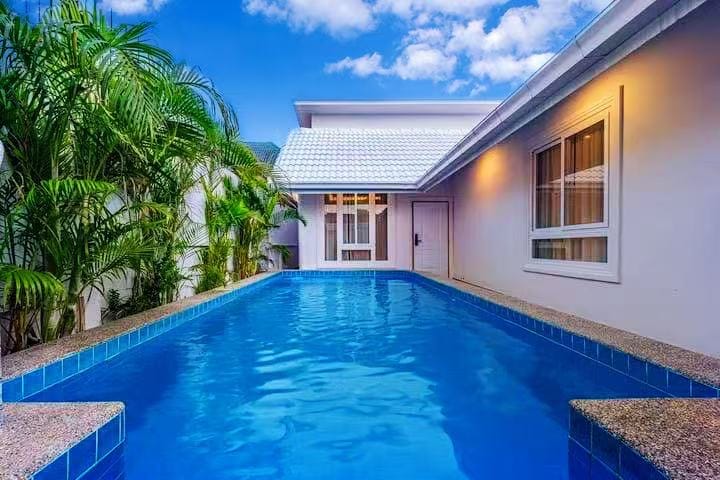 A clear blue swimming pool is set against a backdrop of a light-colored villa. Lush green palm trees are visible alongside the pool, adding a tropical touch. The villa features large windows that enhance the natural light, creating a serene environment.